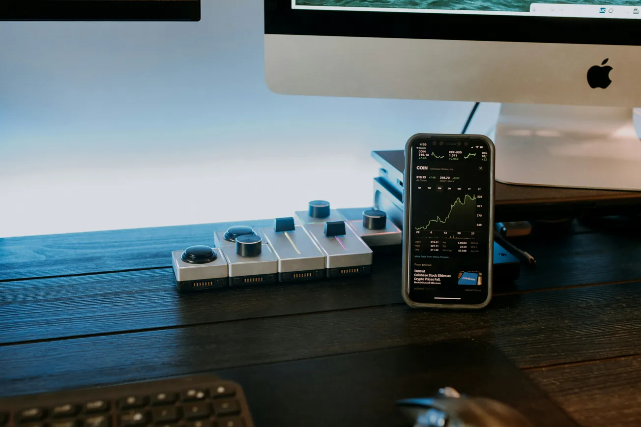 A sleek workspace featuring a smartphone displaying stock market graphs, set against a desktop computer on a dark wooden surface. Tech-savvy and modern ambiance.