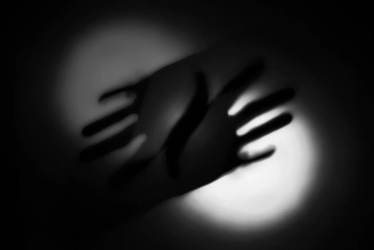 Shadowy silhouette of two hands overlapping against a white light on a dark background, creating an abstract, mysterious, and artistic effect.