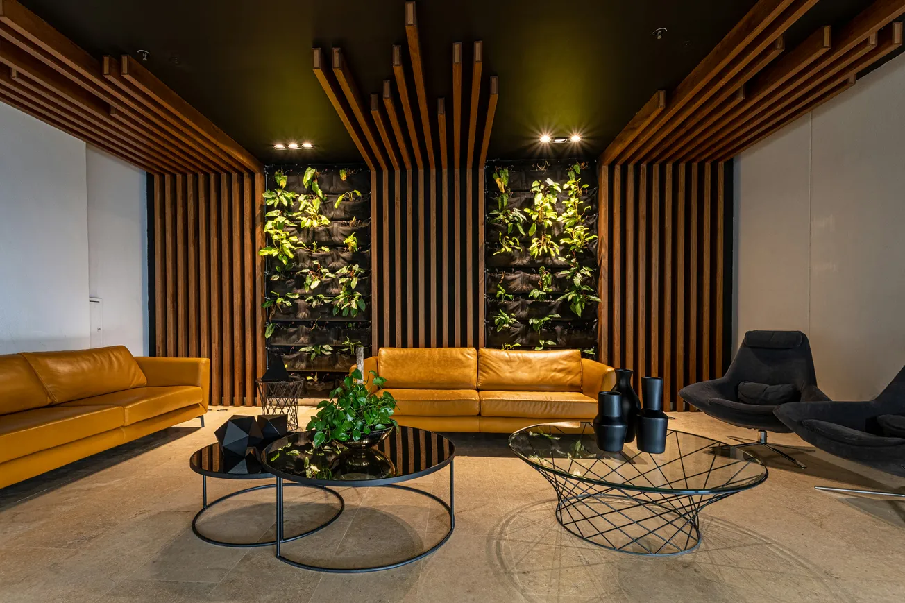 Modern lounge with brown sofas, vertical garden, and wood paneling. Two round coffee tables hold plants and decor, creating a cozy, stylish ambiance.