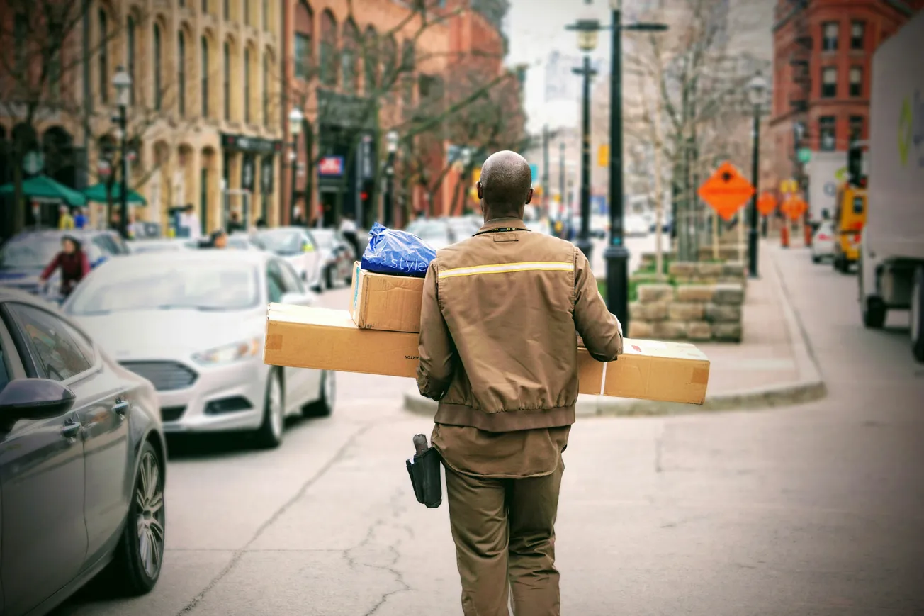 A delivery person in a brown uniform carries packages across a busy city street. Cars line the road, and historic buildings are visible in the background.