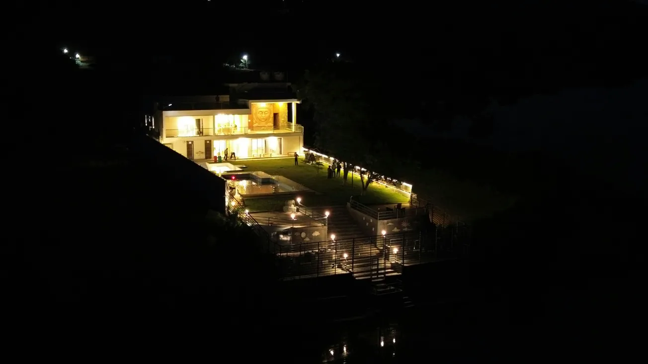Aerial night view of a brightly lit modern house with a spacious lawn. Warm lighting creates a cozy ambiance. Groups of people gather on the lawn.