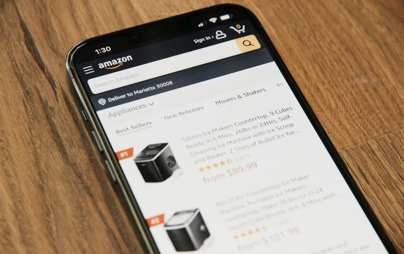 Smartphone displays Amazon shopping app, showing countertop ice makers on a wood surface. Screen displays prices, ratings, and product images.