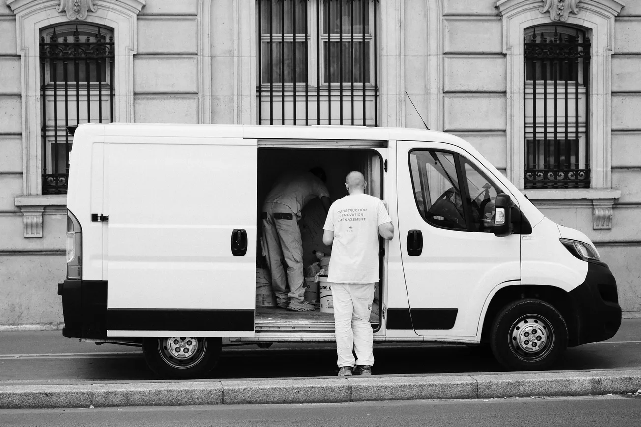 A white van is parked beside a building with ornate windows. Two workers in white uniforms are visible, with one standing outside and the other inside the van.