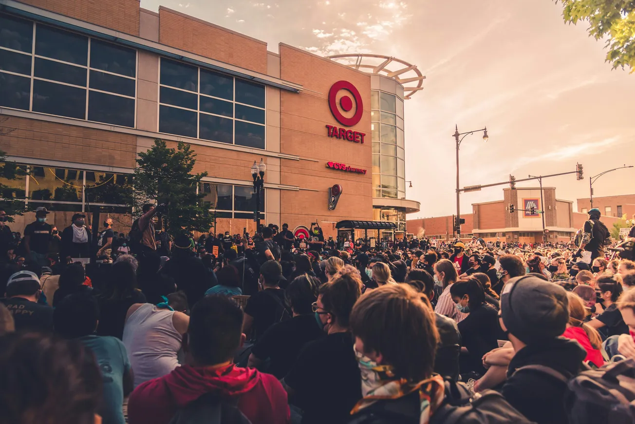 Crowd gathered peacefully outside a Target store during a sunset. People are seated, some wearing masks, conveying solidarity and calm determination.