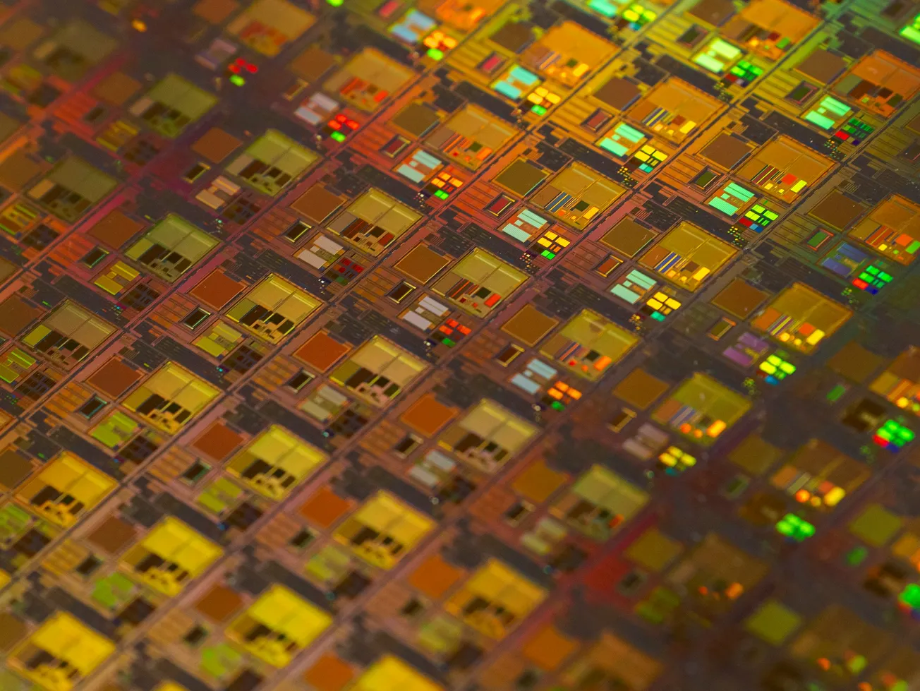 Close-up of a colorful microchip wafer showing intricate square patterns in shades of gold, red, and green, reflecting light with a high-tech feel.