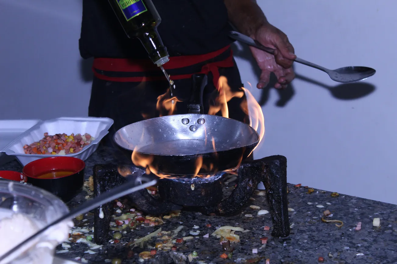 Chef pouring oil into a flaming pan on a stovetop. Nearby, bowls with chopped vegetables and sauce are ready for cooking. Dynamic and intense.