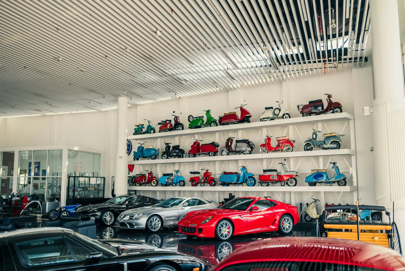 A sleek car showroom with vibrant colors; red sports car in front, flanked by luxury cars. Walls display vintage scooters in various hues, adding retro charm.