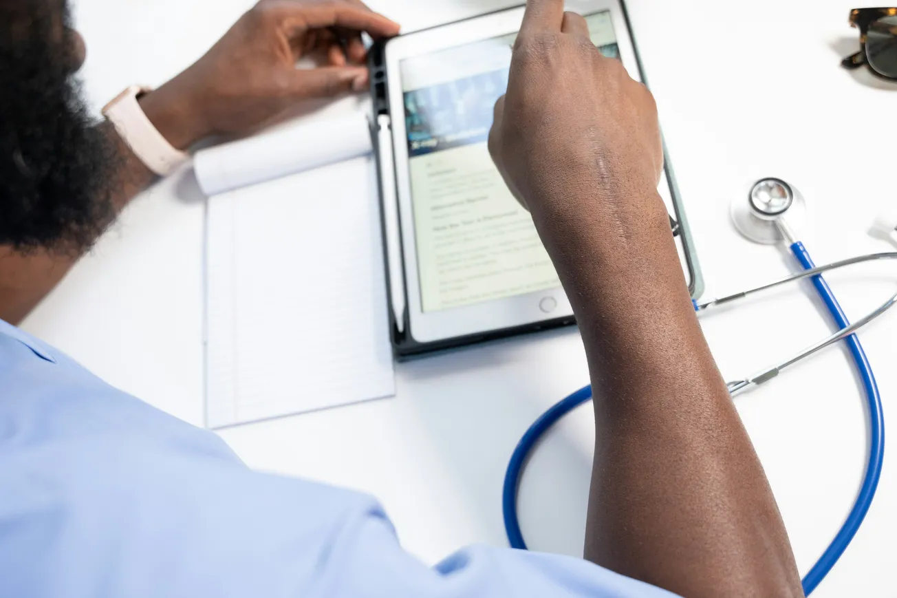 A person in scrubs uses a tablet at a desk with a stethoscope, notebook, and sunglasses. The setting conveys a professional, medical atmosphere.