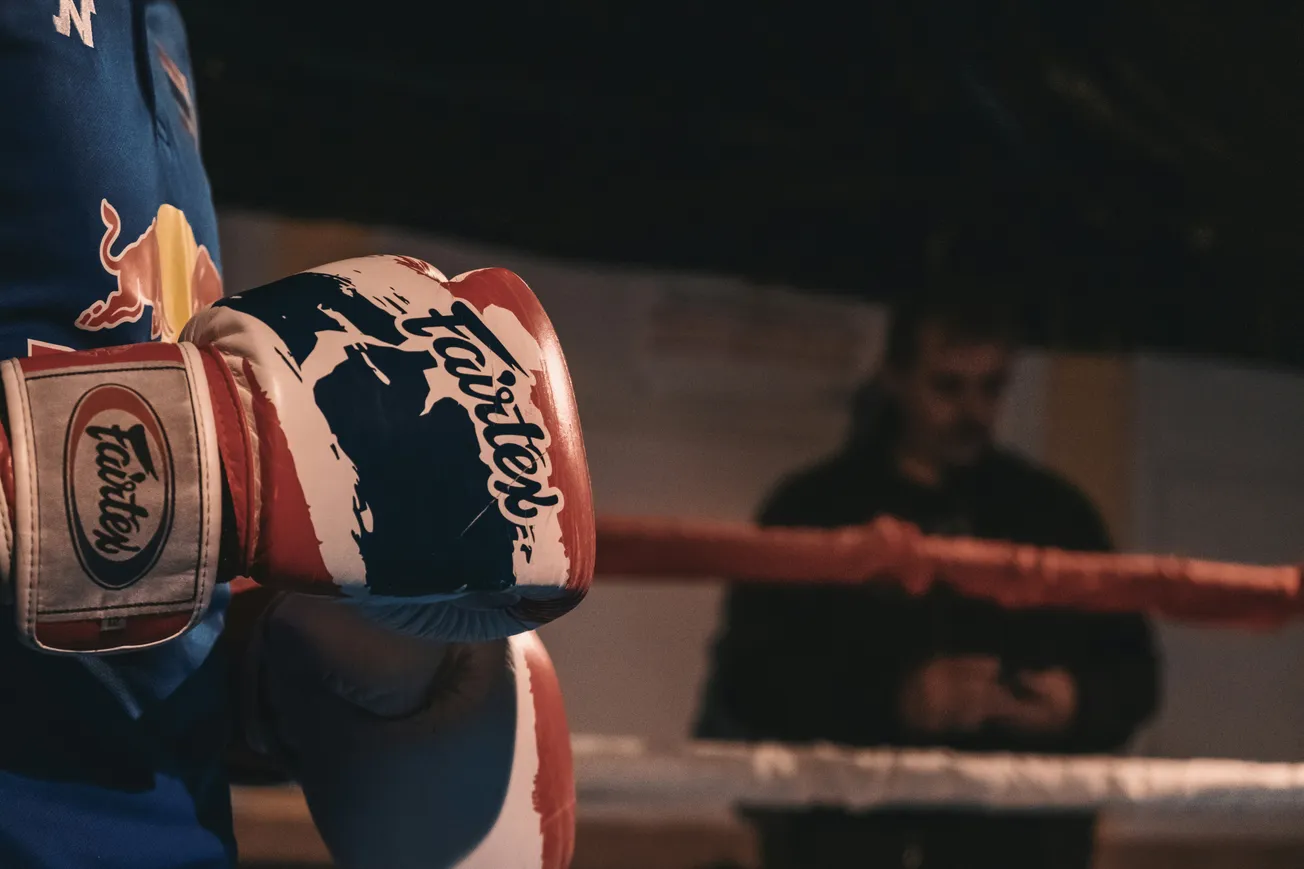 A close-up of a boxer wearing red, white, and blue Fairtex gloves, standing in a dimly lit boxing ring. A person is blurred in the background.