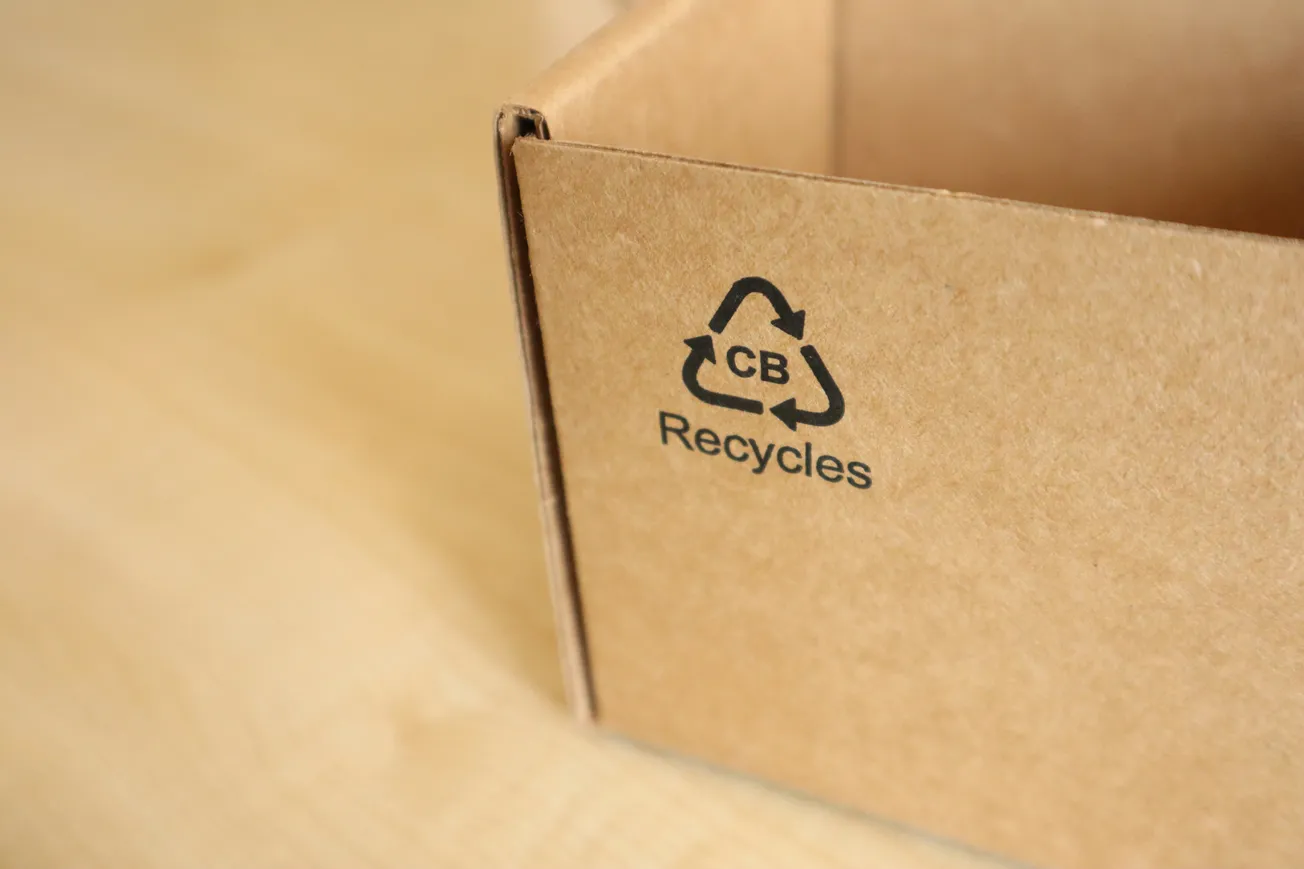Brown cardboard box corner with recycling symbol, "CB Recycles" text. Placed on a light wood surface, symbolizing eco-friendly packaging.
