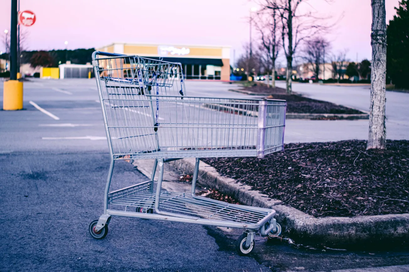 A lone shopping cart sits in an empty parking lot at dusk. The sky is a soft pink, and bare trees surround the area, conveying a peaceful, deserted mood.