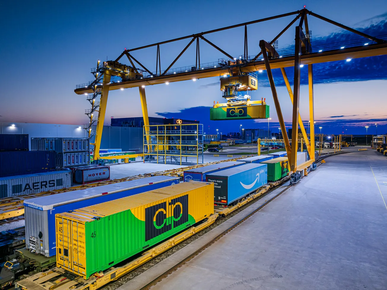 A large crane moves a green shipping container in a well-lit industrial rail yard at dusk. Multiple colorful freight containers line the tracks.