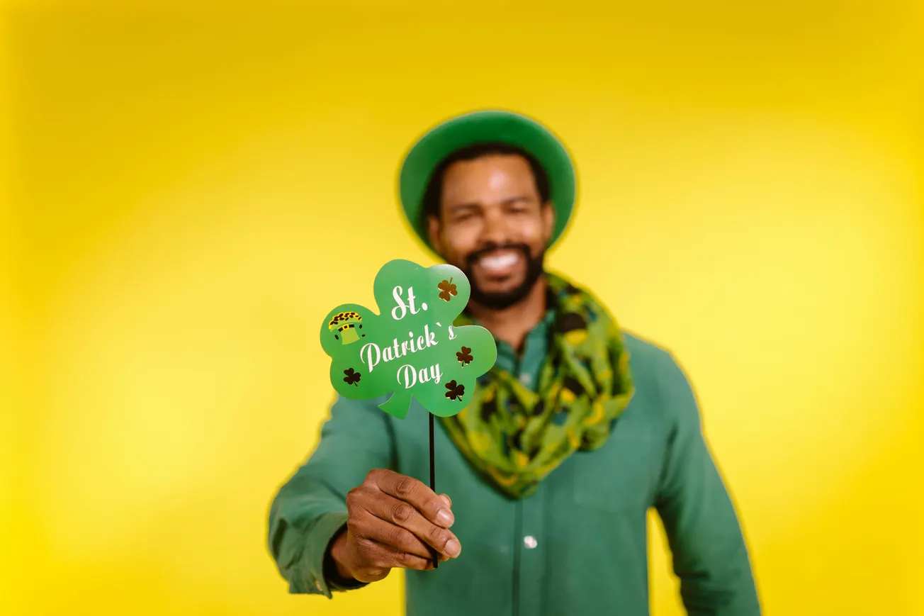 Man in green outfit and hat smiles, holding a green shamrock sign reading "St. Patrick's Day" against a bright yellow background. Festive and cheerful.