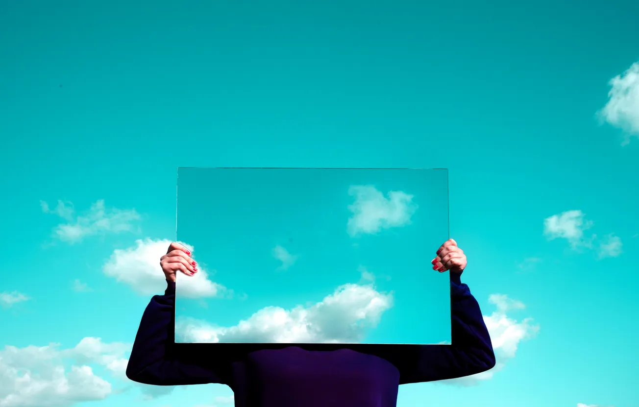 A person holds a rectangular mirror against a bright blue sky with scattered clouds, creating an illusion of a missing head. The scene feels surreal and whimsical.