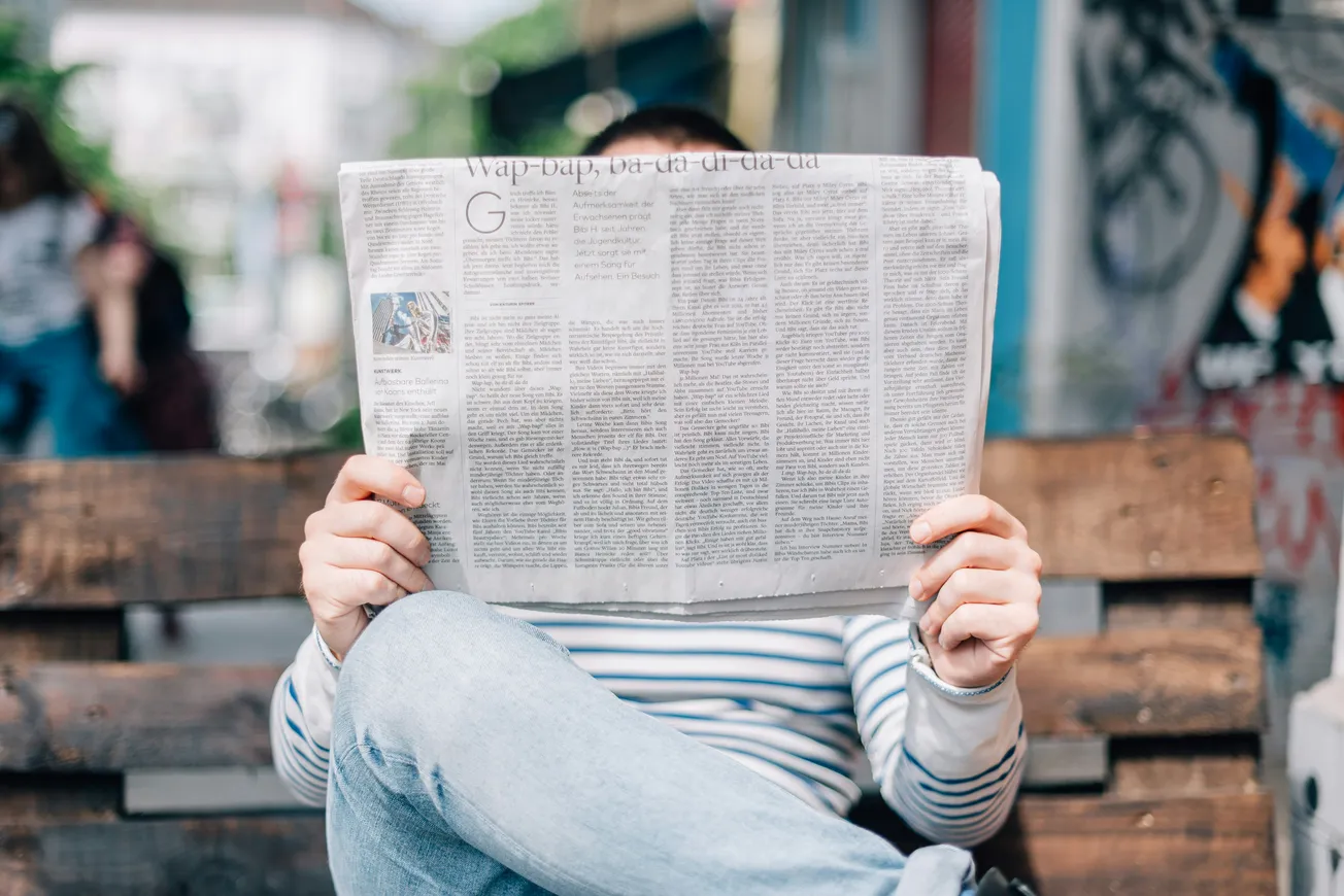 Person sitting on a bench, wearing a striped shirt and jeans, reading a newspaper. The urban background is blurred, conveying a relaxed, casual vibe.