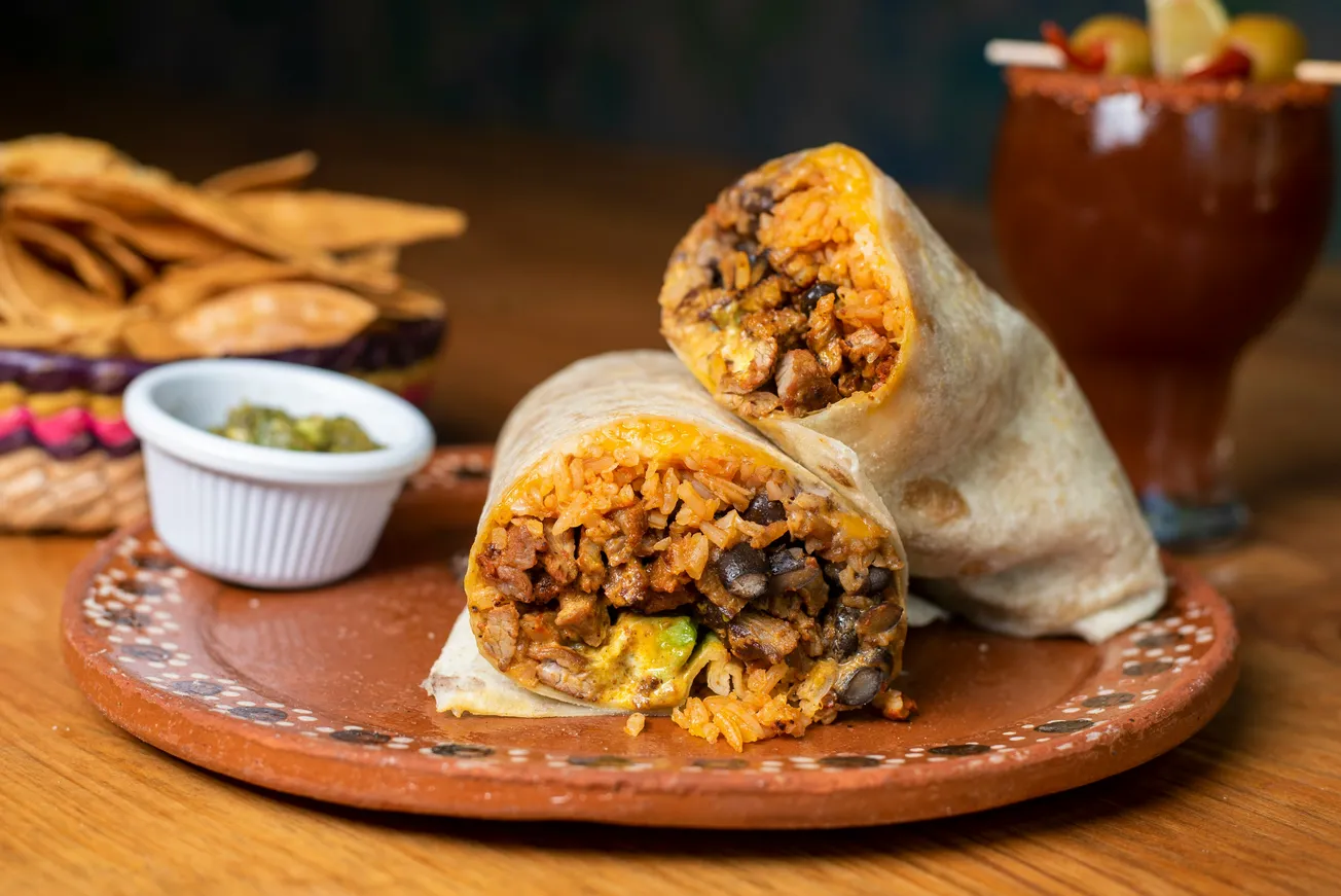 A delicious burrito, halved to reveal rice, beans, and mixed fillings, sits on a rustic plate. Chips, salsa, and a cocktail are blurred in the background.