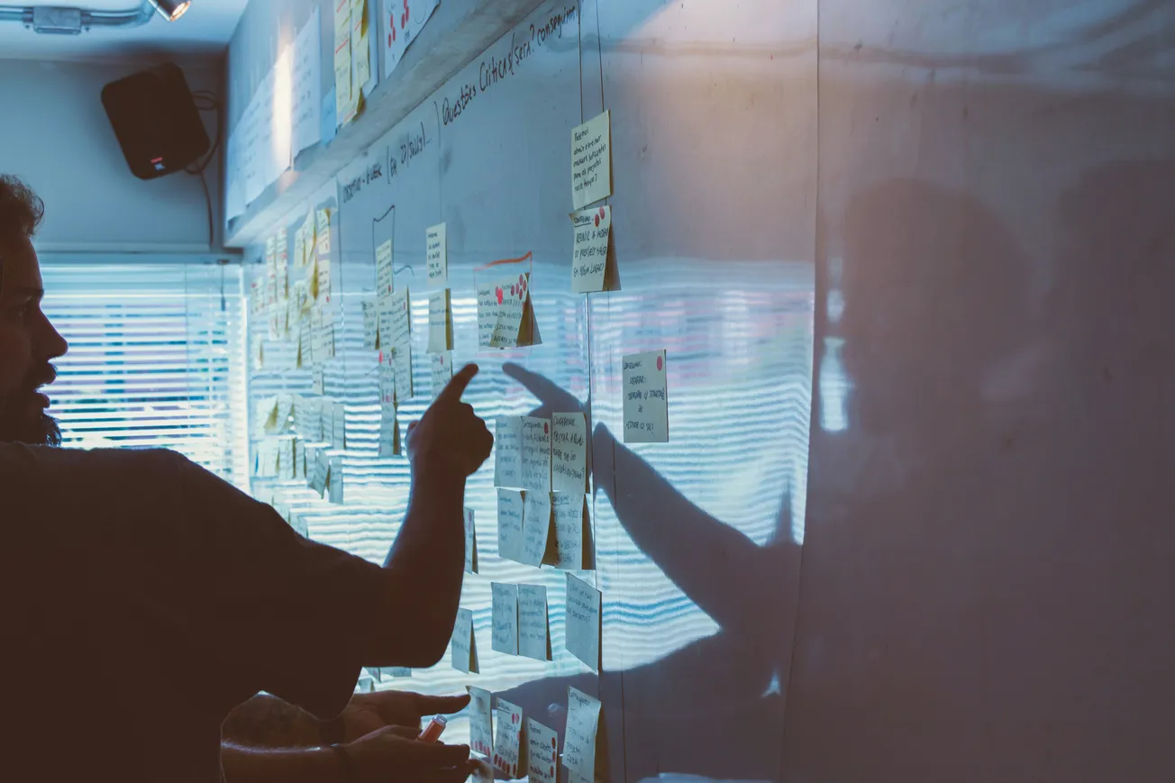 A person gestures toward sticky notes on a wall in a dimly lit room, suggesting a collaborative and focused environment. Shadows add depth and intensity.