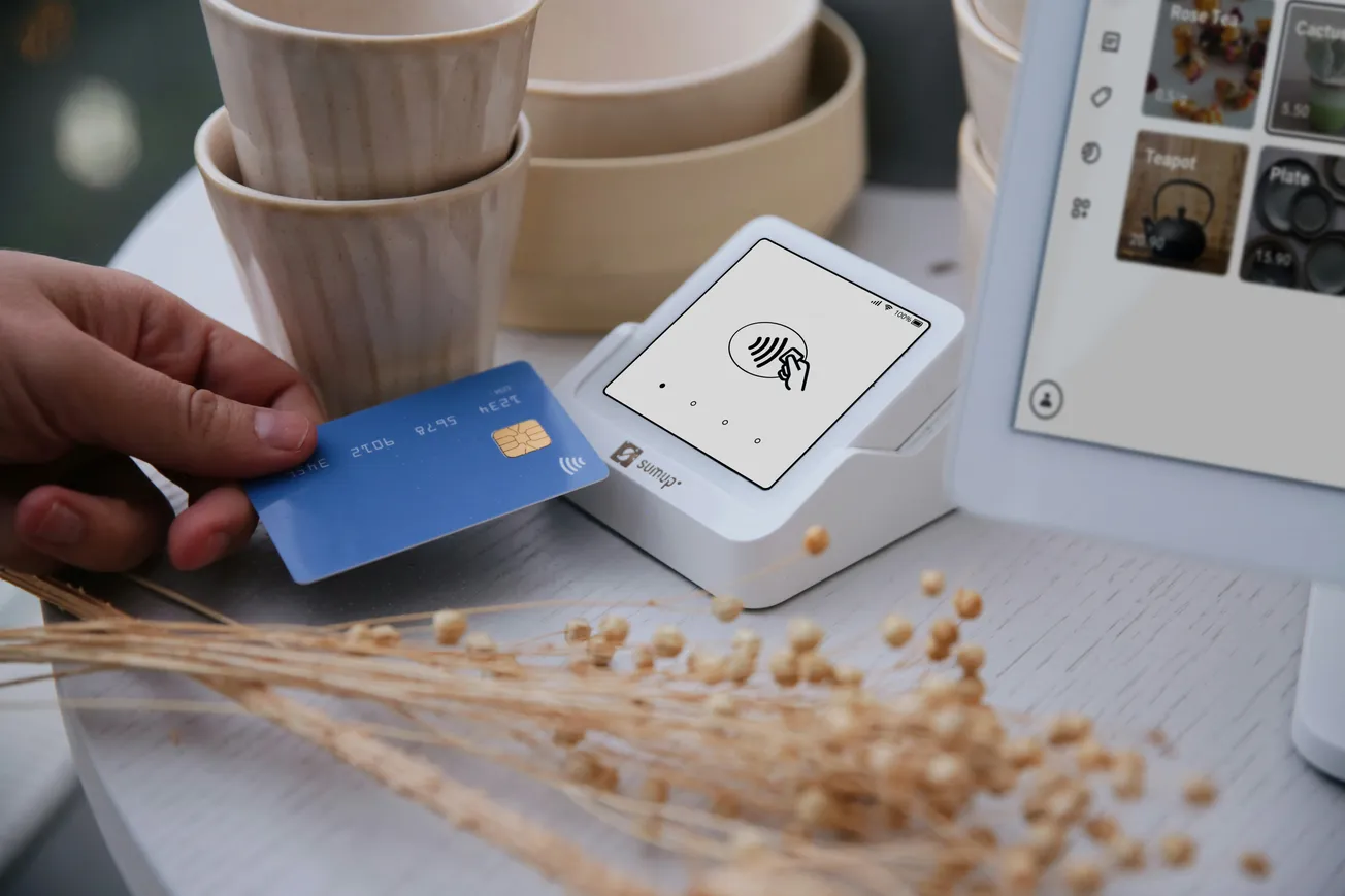 A person taps a blue contactless credit card on a white payment terminal. In the background, there are stacked cups and a tablet screen displaying product images.