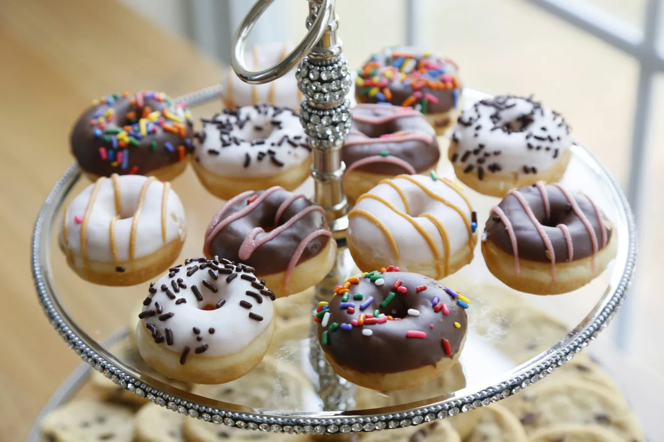 A silver tiered tray holds an assortment of mini donuts with colorful toppings. Frostings include white and chocolate with sprinkles and drizzles. Bright and inviting.