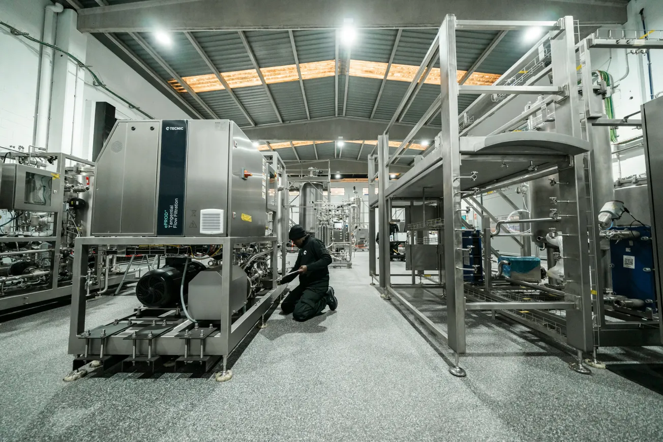 A worker in dark clothing inspects industrial machinery in a spacious, well-lit factory. The scene conveys a sense of focus and precision.