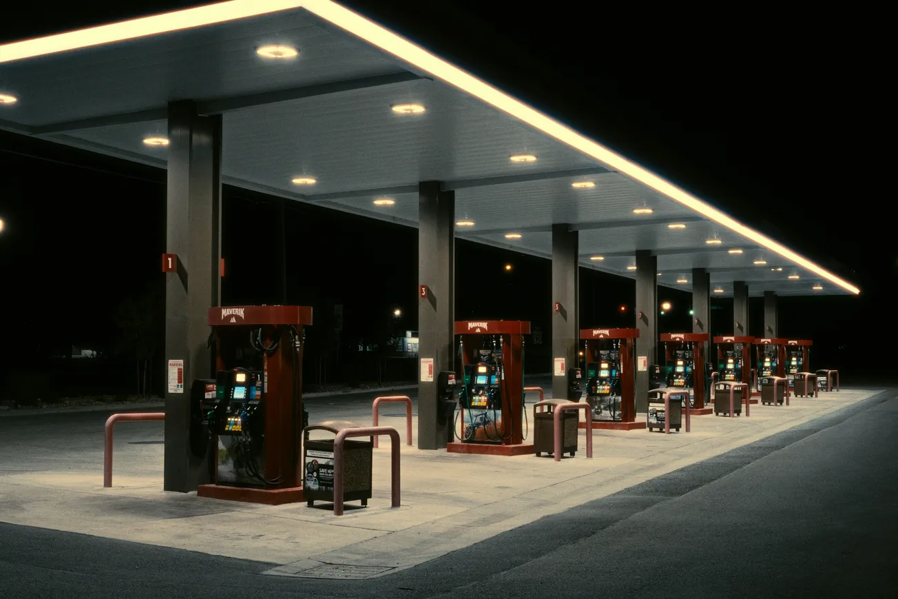 A brightly lit gas station at night, featuring a row of red fuel pumps under a wide canopy. The area is empty, creating a quiet and isolated atmosphere.