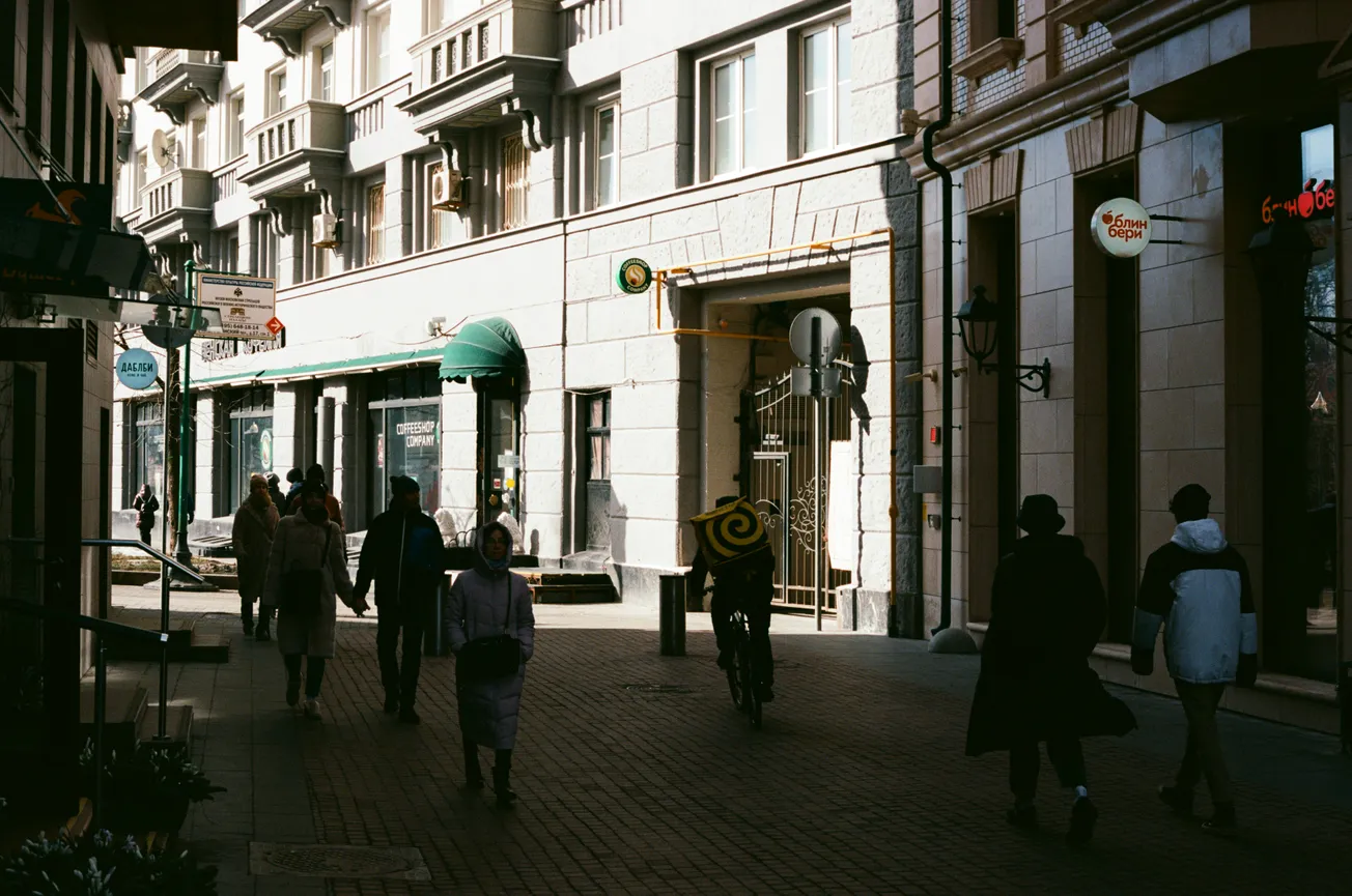 A sunlit urban street with people walking, some wearing coats. A cyclist with a yellow-black spiral backpack rides by. Buildings feature ornate facades.