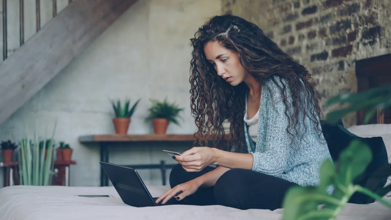 A woman with curly hair sits on a bed, intently looking at a laptop while holding a credit card. Surrounding her are green plants, creating a cozy atmosphere.