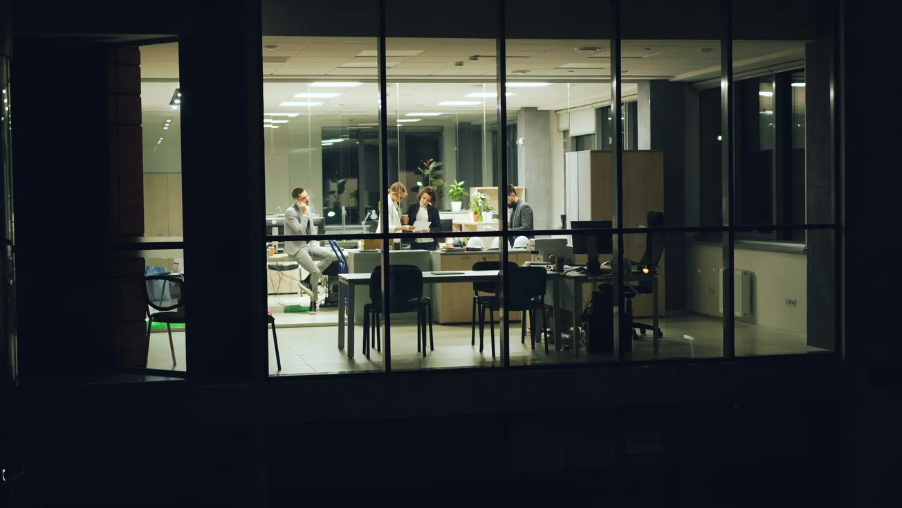 A small group of people are engaged in a nighttime office meeting, visible through large windows. The space is brightly lit, conveying a focused atmosphere.