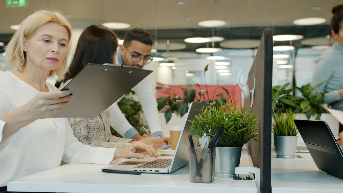 An office setting with people working at desks. A woman in focus examines a clipboard, while colleagues collaborate in the background. Green plants add vibrancy.