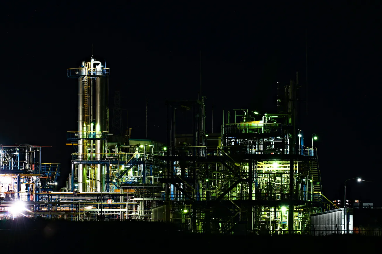 Illuminated industrial plant at night with a complex network of pipes and towers. Bright lights highlight the metal structures against the dark sky.