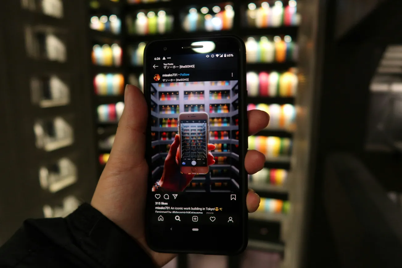 A hand holds a smartphone displaying an Instagram post, capturing a similar image of colorful bottles on shelves in the background, creating a visual loop effect.