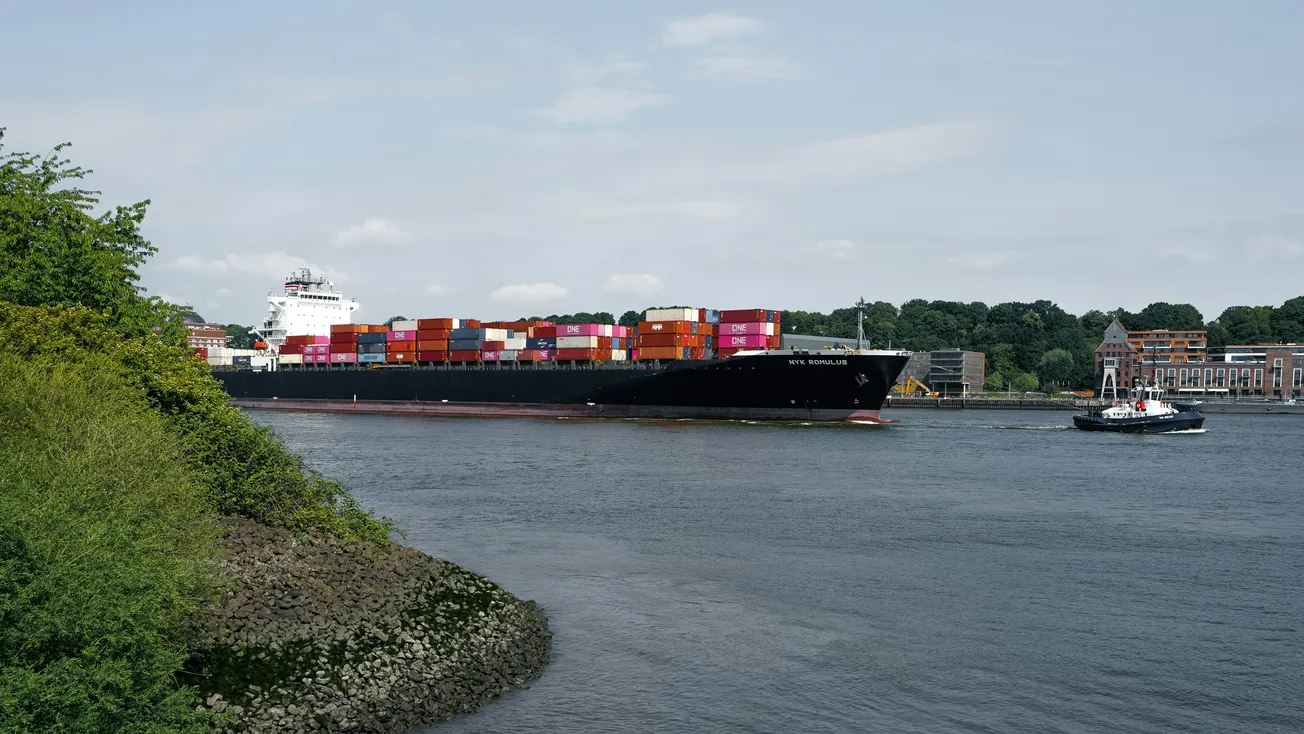 A large cargo ship loaded with colorful containers travels along a calm river, accompanied by a smaller tugboat. Lush greenery and buildings line the riverbank.