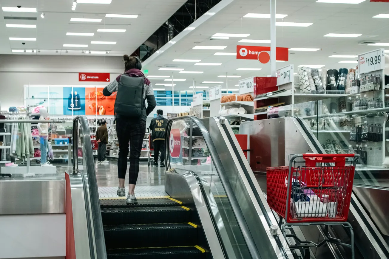A woman with a backpack ascends an escalator in a brightly lit department store. A red shopping cart is nearby, and shelves display various home goods.