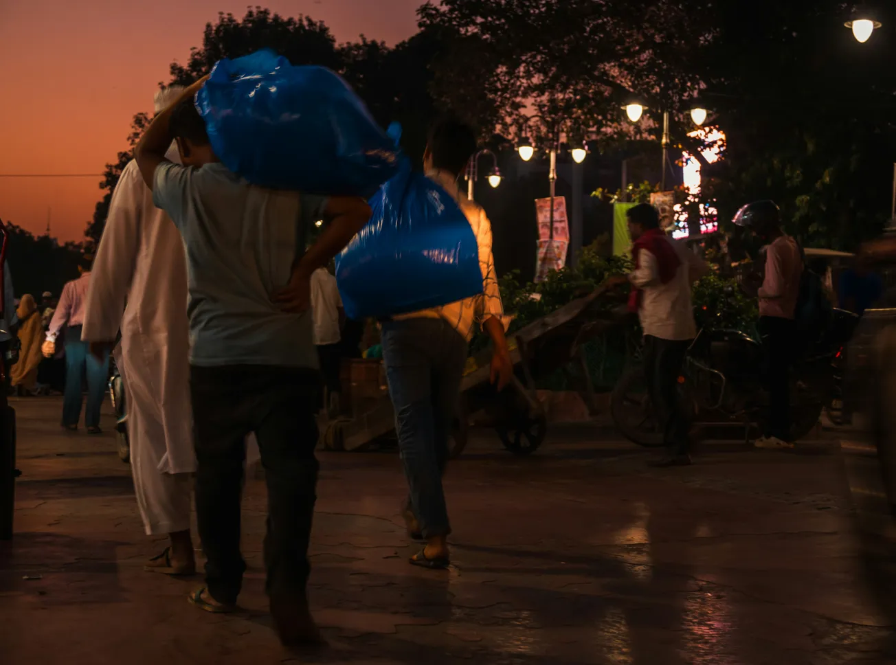 A busy street scene at dusk. People walk under glowing streetlights, one carrying a large blue bag. The atmosphere is lively and bustling.