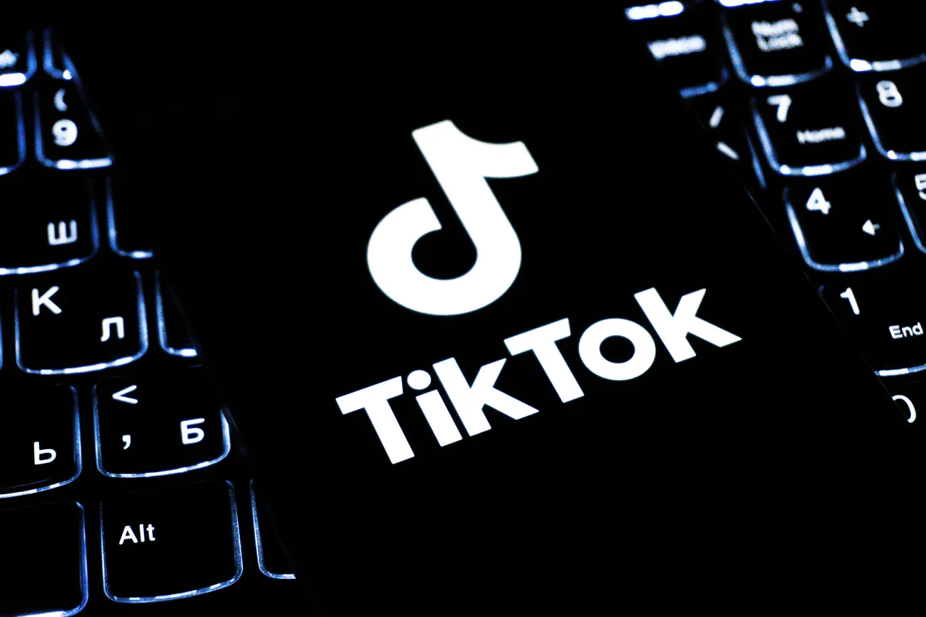 A smartphone displaying the TikTok logo rests on a dimly lit keyboard. The glow from the screen and keys creates a tech-focused, modern ambiance.